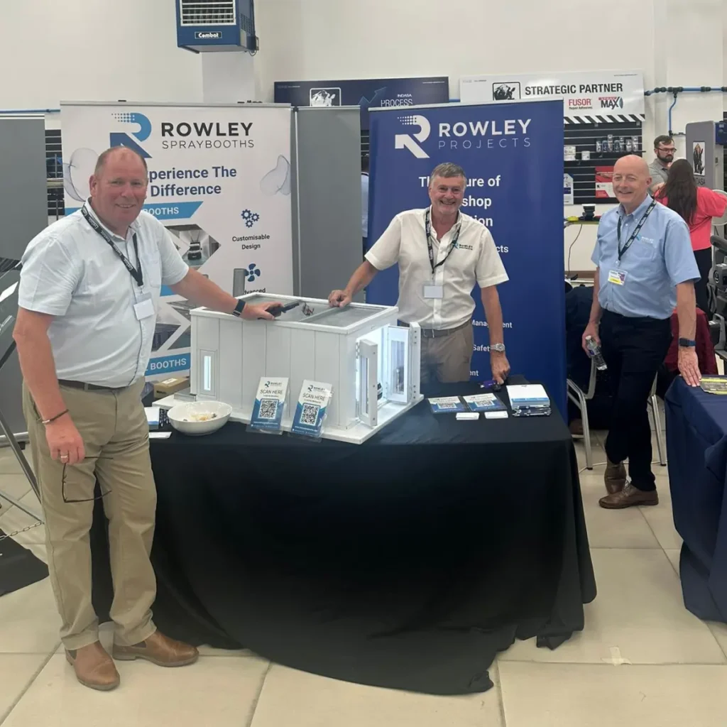 Three men stand around a display stand for Rowley Projects at an indoor event. The stand features banners, brochures, a small display model, and a bowl of sweets on the table. Other stands and people are visible in the background.